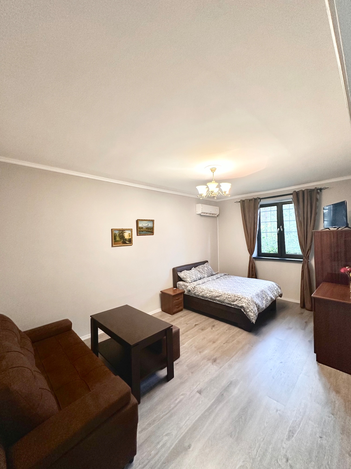 A cheerful living space is depicted, featuring a spacious bed adorned with light bedding, accompanied by a small bedside table. A brown sofa and a wooden coffee table occupy the area. Natural light enters through a window, and soft curtains frame the view.