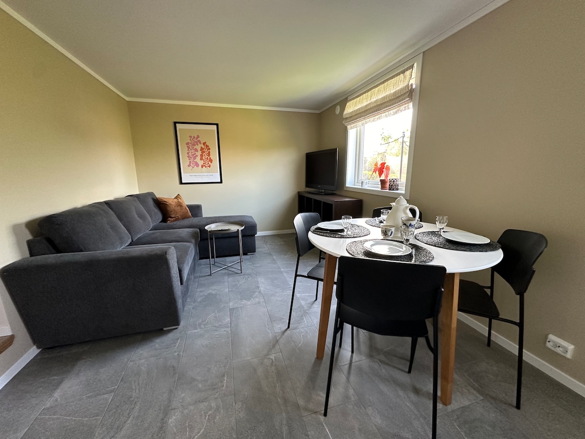 A spacious living area features a sectional sofa and a dining table set for four. Natural light enters through a window, illuminating the grey tile floor. A television is positioned in the corner, providing a cozy atmosphere for relaxation.