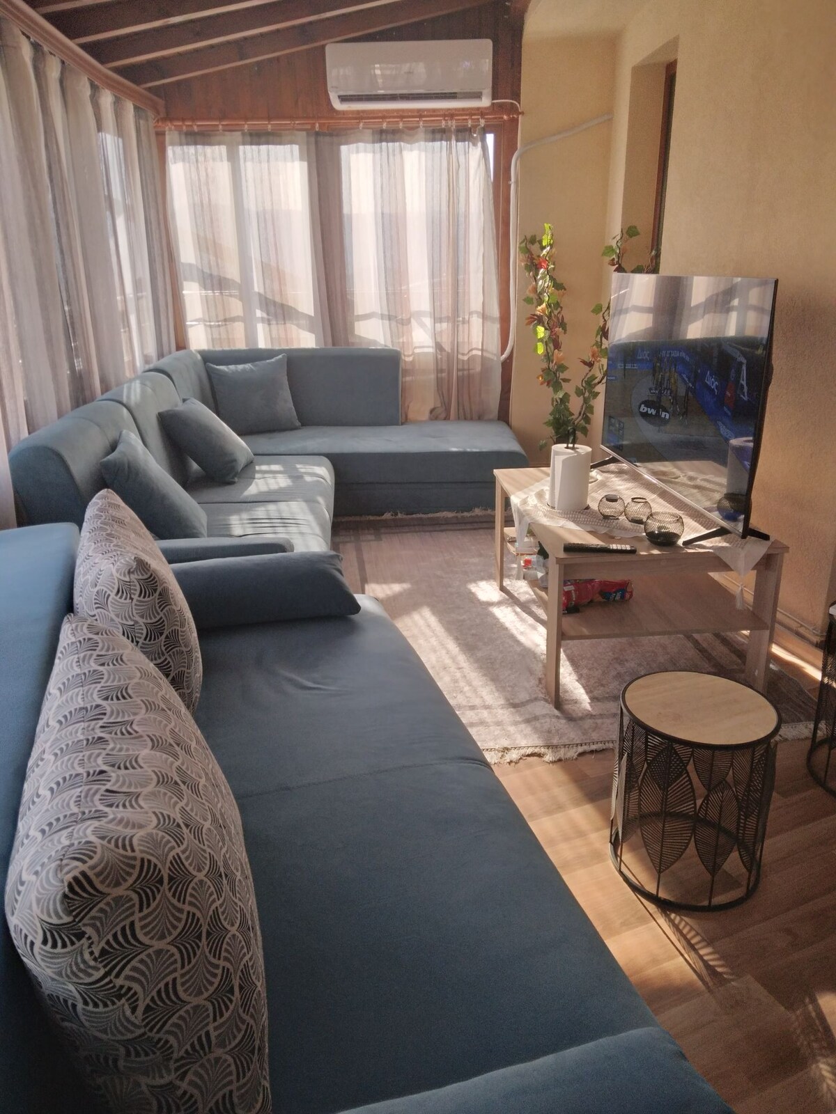 A spacious living area is presented with light blue sofas and decorative cushions, arranged around a central coffee table. A large television is positioned on a stand opposite the seating, while sunlight filters through sheer curtains, creating a bright and airy atmosphere.