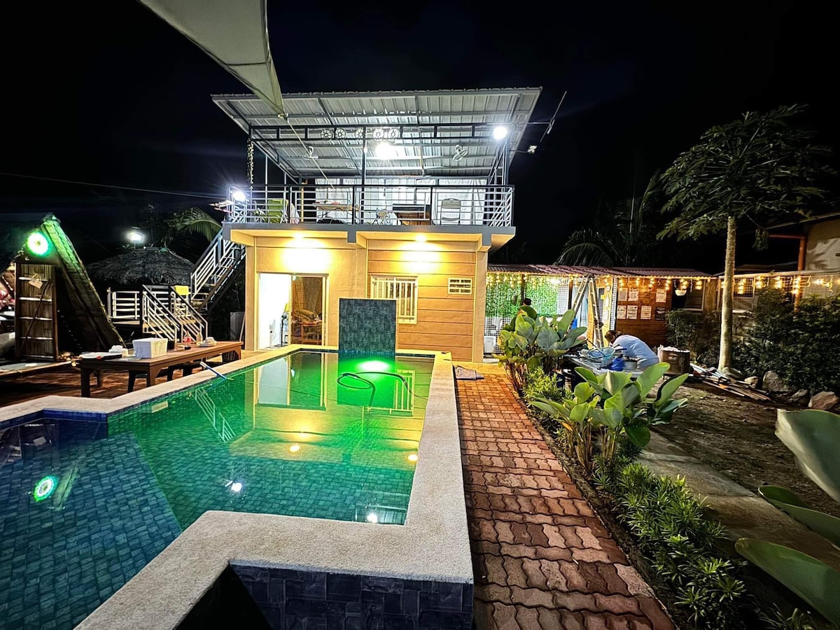A modern outdoor space features a swimming pool illuminated with green lighting. A two-story building is visible in the background, with exterior lights highlighting its structure. The surrounding garden includes well-maintained plants and a paved pathway, creating a serene ambiance at night.