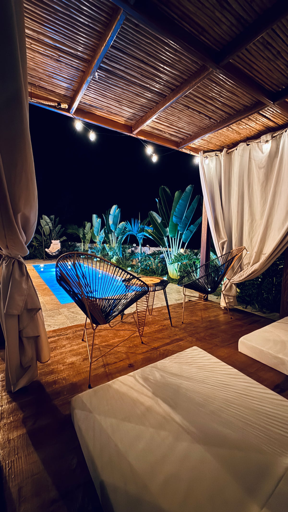 An outdoor seating area features two stylish chairs beside a table, under a covered patio. A pool is visible in the background, surrounded by lush greenery and softly glowing string lights, creating an inviting nighttime ambiance.