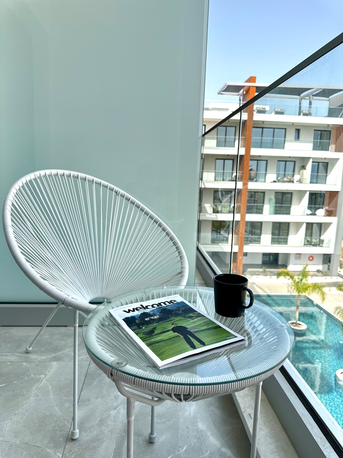 A cozy seating area features a white chair and a round table. A magazine titled 'Welcome' rests on the table, accompanied by a black mug. Large glass windows provide a view of a landscaped pool area and nearby buildings.