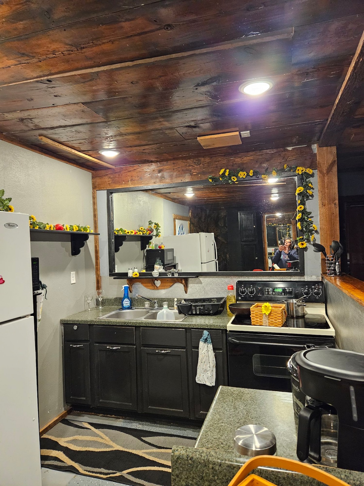 A well-equipped kitchen features dark cabinetry and a contrasting light granite countertop. A refrigerator and stove are visible, along with small appliances and utensils. Decorative elements, including yellow flowers, add a cheerful touch. Warm lighting enhances the inviting atmosphere in the space.