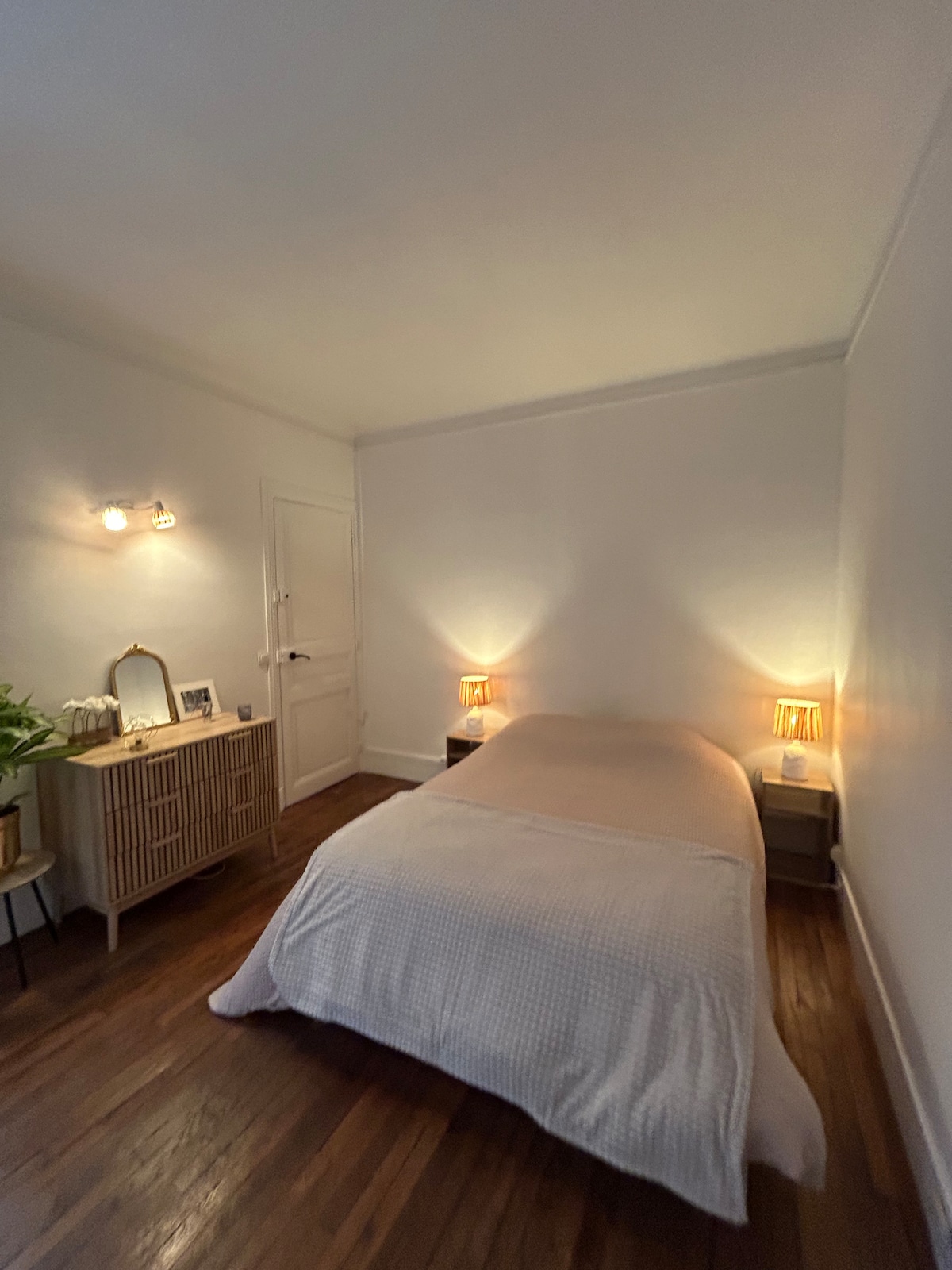 A cozy bedroom features a single bed with a soft, light-colored bedspread, complemented by bedside tables with warm lamps. A mirror is positioned on a dresser, and a decorative plant adds a touch of greenery. The space is illuminated by soft lighting, creating a tranquil ambiance.