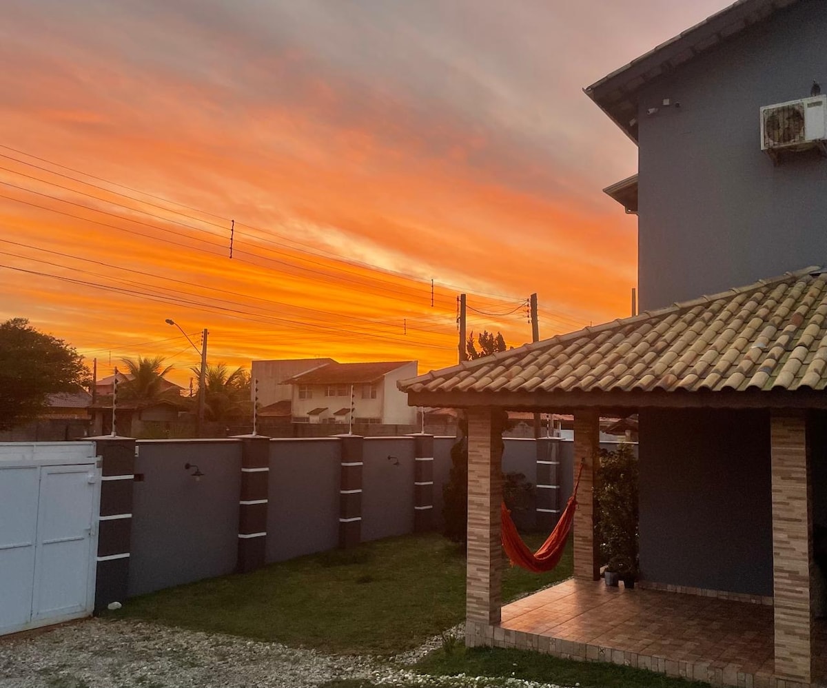A charming outdoor view features a vibrant sunset sky painted in hues of orange and pink. A shaded patio area with a traditional roof provides a relaxing space, while a hammock is visible, inviting calm moments. Lush grass and a gravel path enhance the setting.