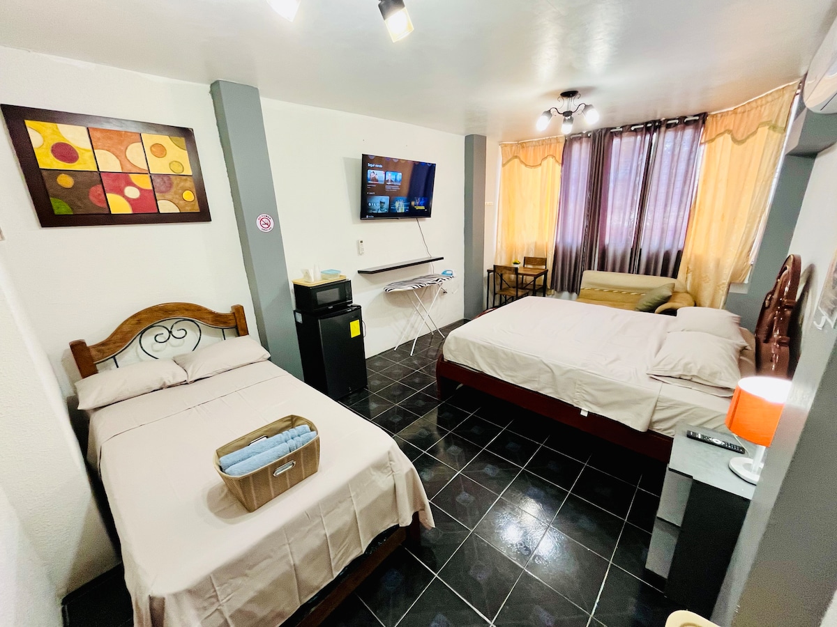 A cozy suite is shown, featuring two beds with neutral bedding and a wooden headboard. A flat-screen TV is mounted on the wall. Natural light filters through sheer curtains, illuminating a small seating area with a sofa. An iron and board are positioned nearby.