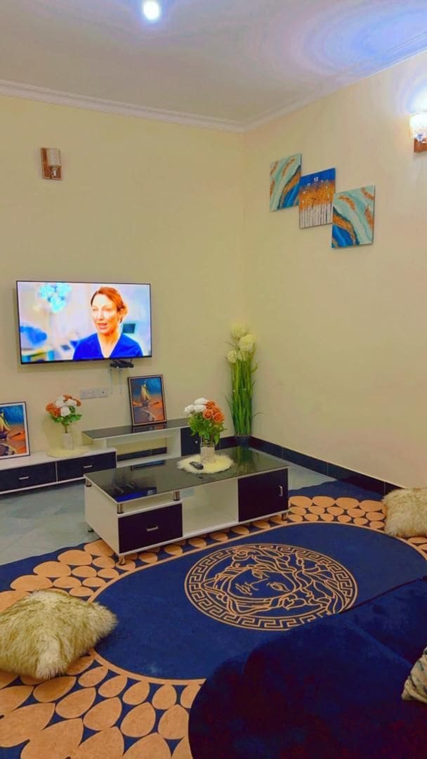 A cozy living area is presented with a dark blue circular rug and plush seating arrangements. A modern TV is mounted on the wall above a low coffee table, which is accompanied by decorative plants and floral arrangements. Bright wall art adds a touch of color.