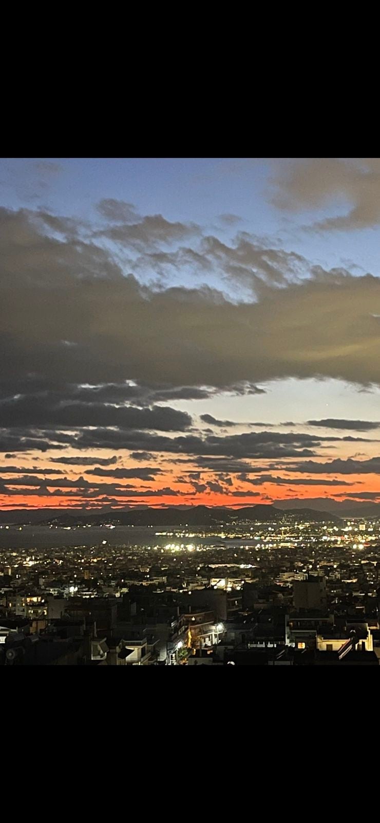 A panoramic view of the city skyline is captured during sunset, showcasing rich hues of orange, blue, and gray in the sky. The twinkling city lights below contrast beautifully against the dramatic clouds, creating a serene evening atmosphere.