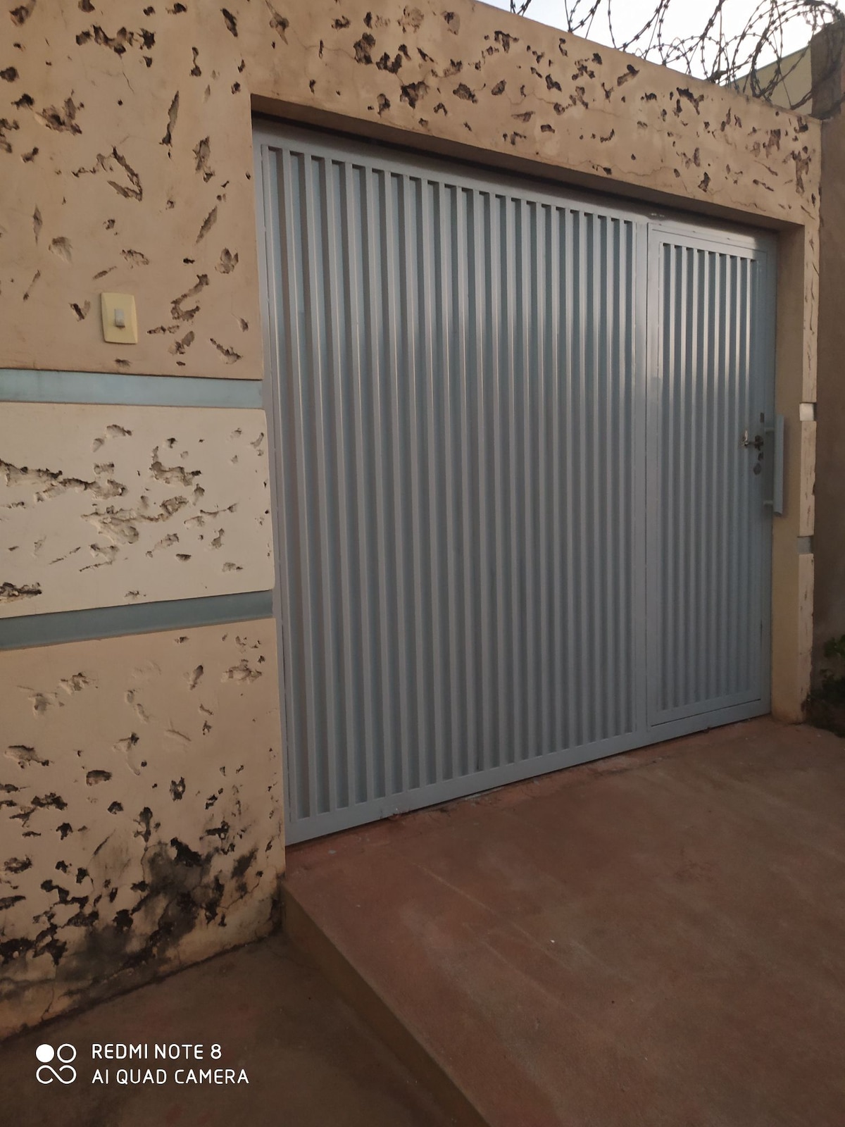 An outdoor entrance features a light blue metal gate, secured with a latch on the right side. Textured walls surround the gate, displaying a worn and rustic appearance. A concrete pathway leads up to the entrance, providing access to the property.