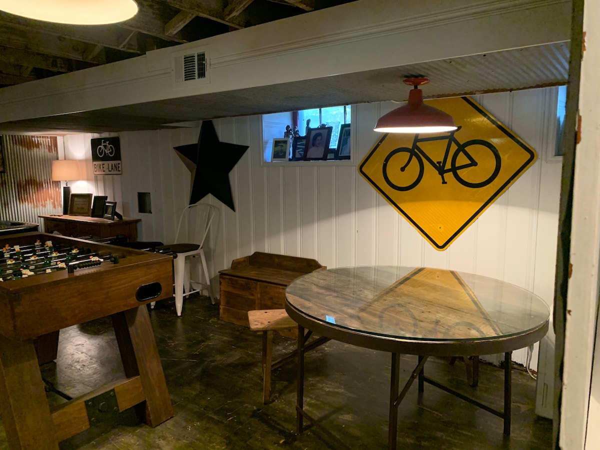 A basement entertainment area includes a round table with a glass top and wooden chairs, creating a casual dining space. A foosball table stands nearby, while a large bicycle-themed sign and a star decoration adorn the wall, adding character to the environment.