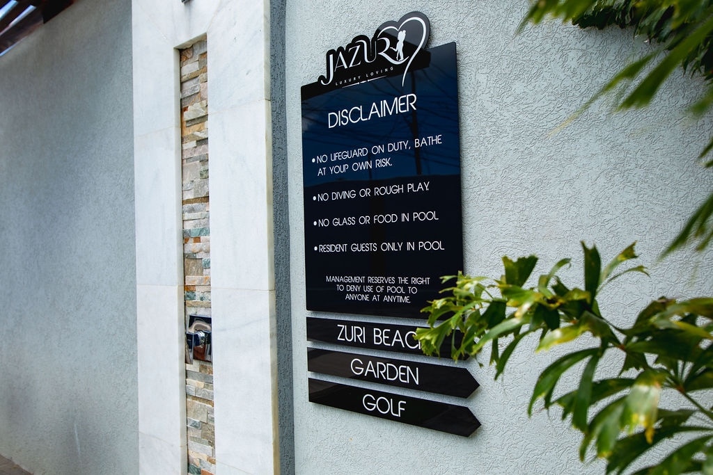 A sleek and modern signage board is displayed on a textured wall. The board features clear white lettering providing guidelines and disclaimers for guests, surrounded by greenery that adds a touch of nature to the setting.