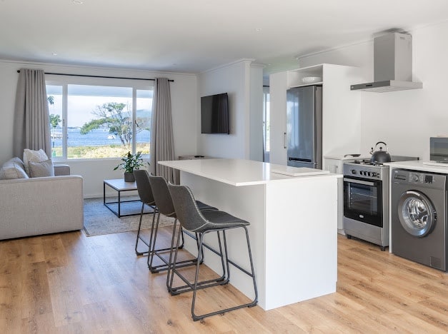 An open-plan kitchen features modern appliances, including a washing machine and oven. Bar stools line a sleek white countertop. Natural light floods the space through large windows with scenic views of the lagoon, and a cozy seating area with a sofa is nearby.