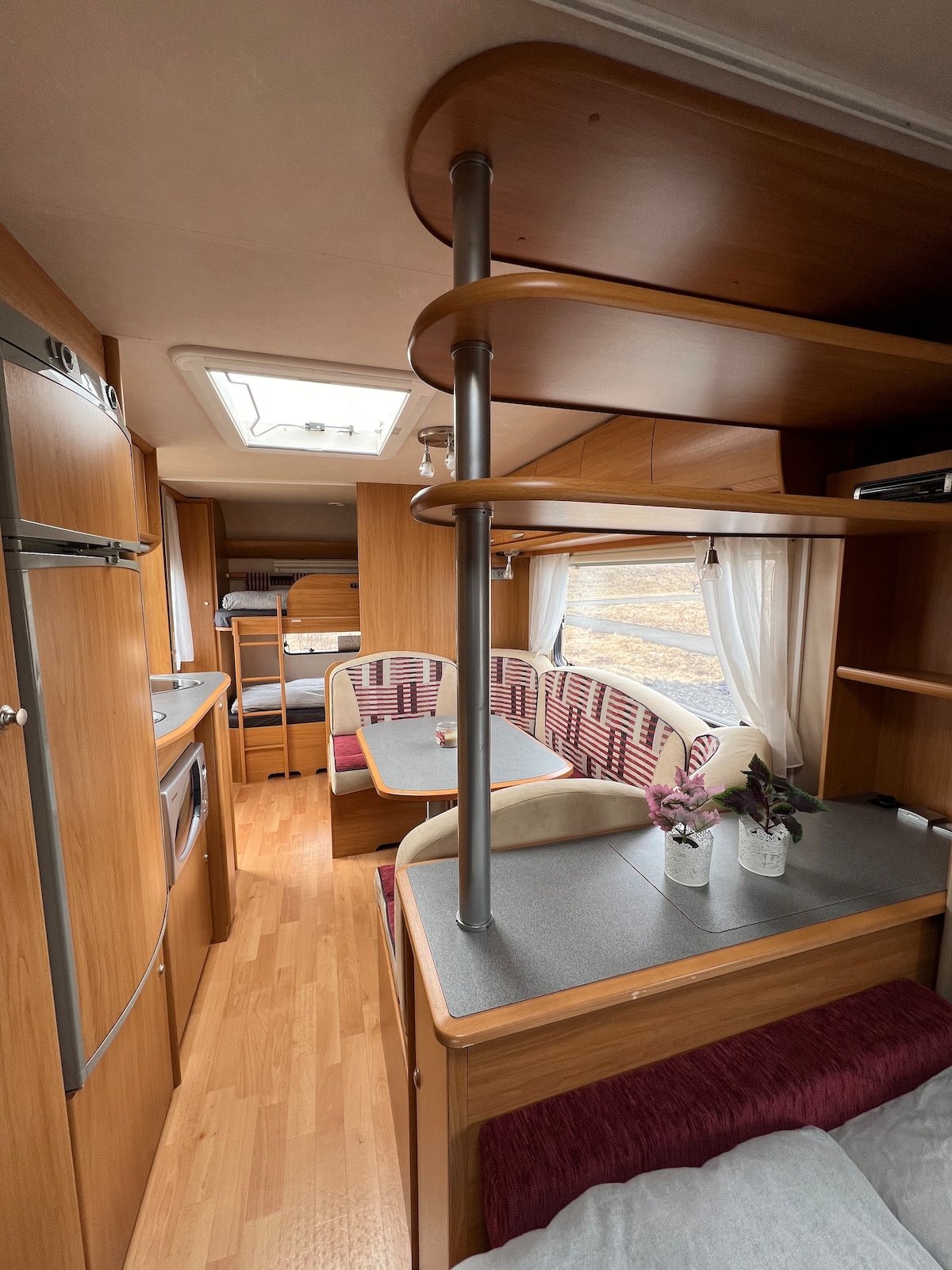 The interior of the camper home features wooden cabinetry and a functional layout. A dining area is complemented by a table surrounded by upholstered seating, while the kitchen area includes essential appliances. Natural light enters through a skylight, enhancing the warm wood tones.