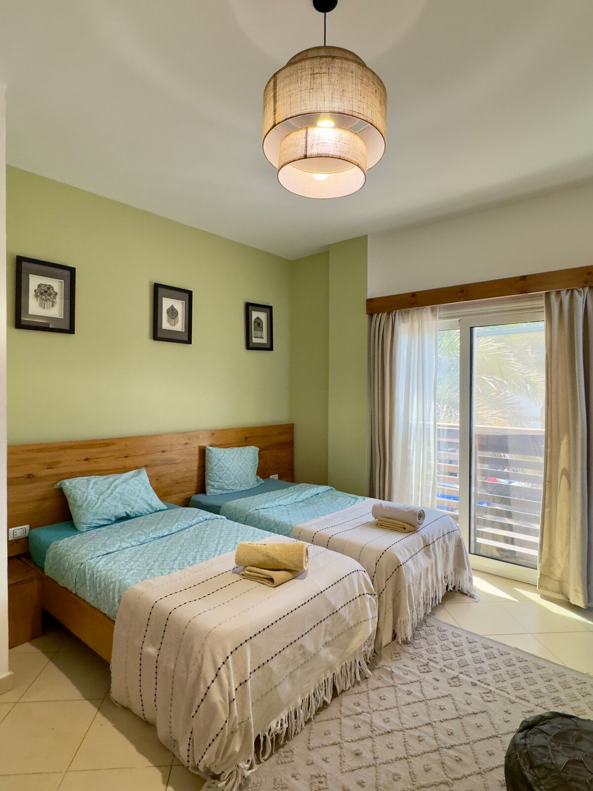 A double room featuring two spacious beds dressed in light blue covers, complemented by soft blankets. Natural light enters through sliding glass doors leading to a small balcony. Framed art is displayed on the green walls, contributing to a calming atmosphere.