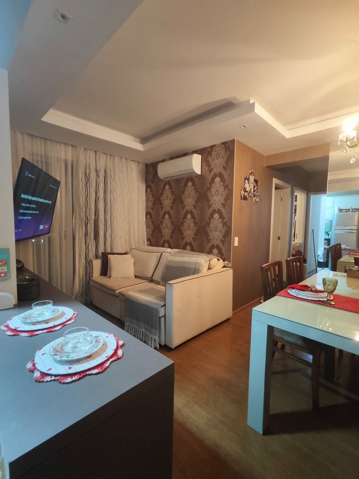 An inviting living area features a light-colored sofa adorned with soft pillows, set against a stylish patterned wall. A modern TV is mounted on the opposite wall, while a dining table with chairs is positioned nearby. Natural light filters through sheer curtains.