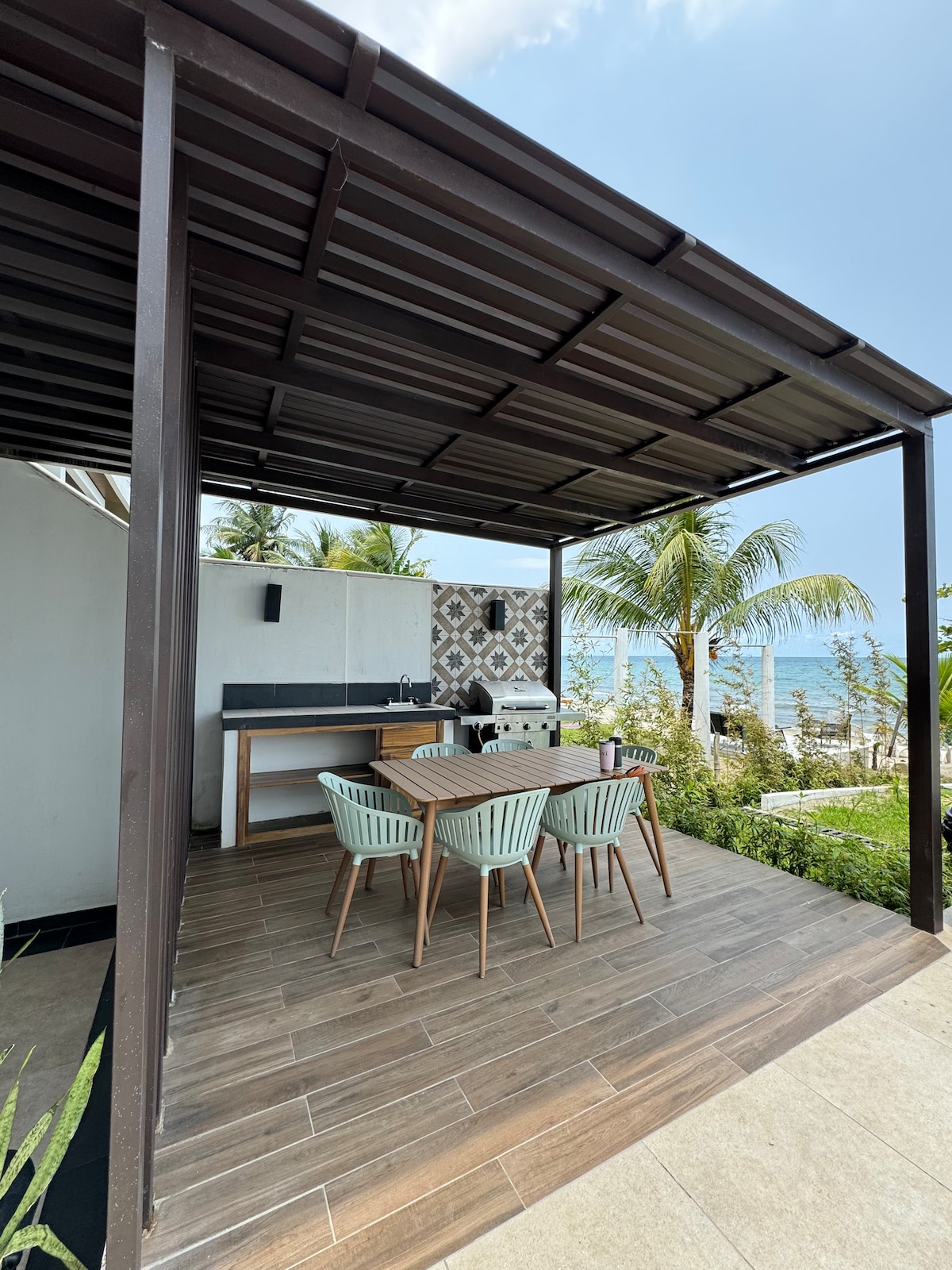 An outdoor dining space features a sturdy wooden table surrounded by four light green chairs. A minimalist bar counter is visible in the background, complemented by a decorative wall. Lush greenery and palm trees frame the setting, while the ocean is visible in the distance.