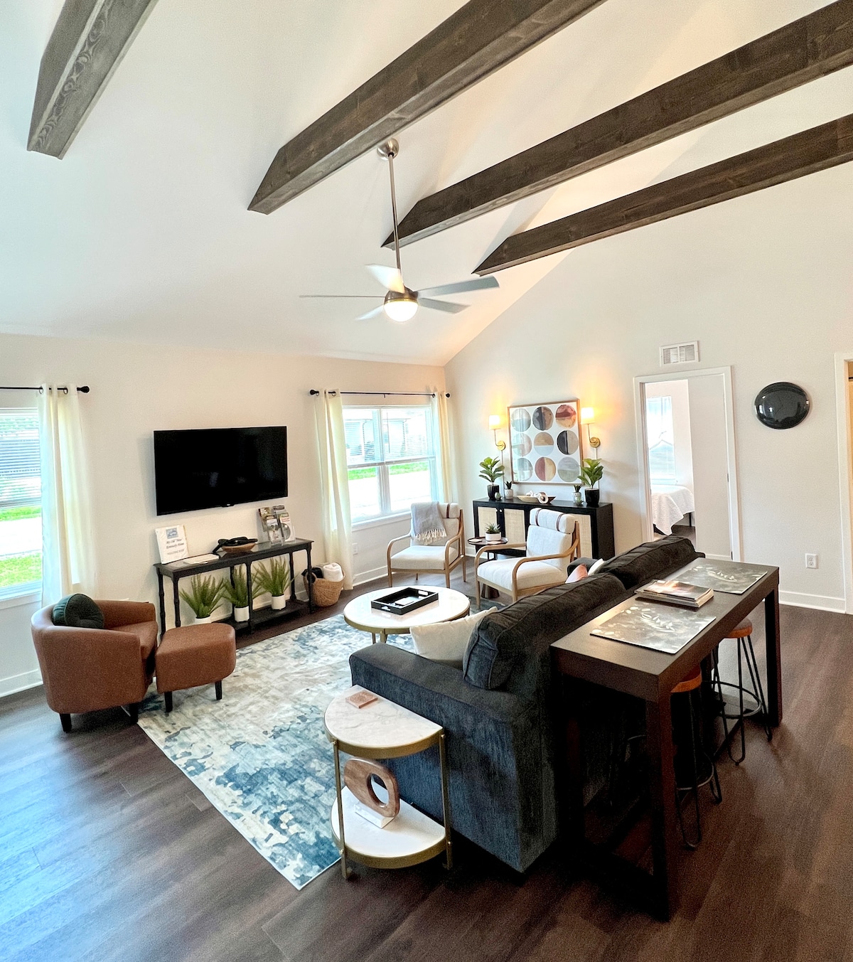A spacious living area features a cozy sectional couch facing a wall-mounted TV. Natural light enters through large windows, illuminating the modern decor. Distinctive ceiling beams are highlighted, and a stylish area rug grounds the space, complemented by a small dining table and two armchairs.