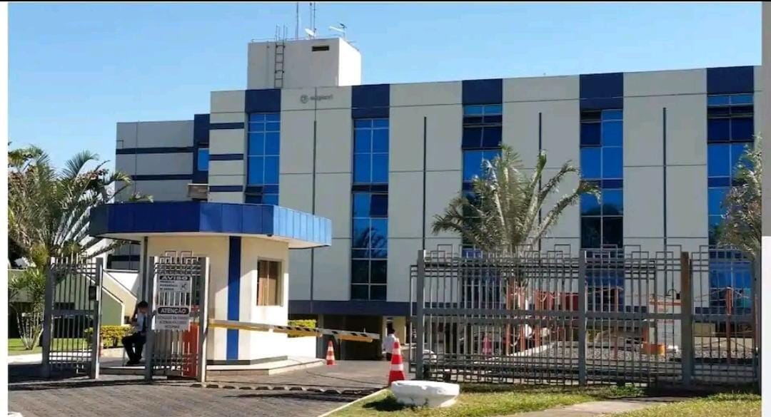 A modern building is shown with a blue and white facade. The entrance is gated, featuring a security booth and palm trees lining the pathway. Large windows reflect the clear sky, enhancing the welcoming appearance of the establishment.