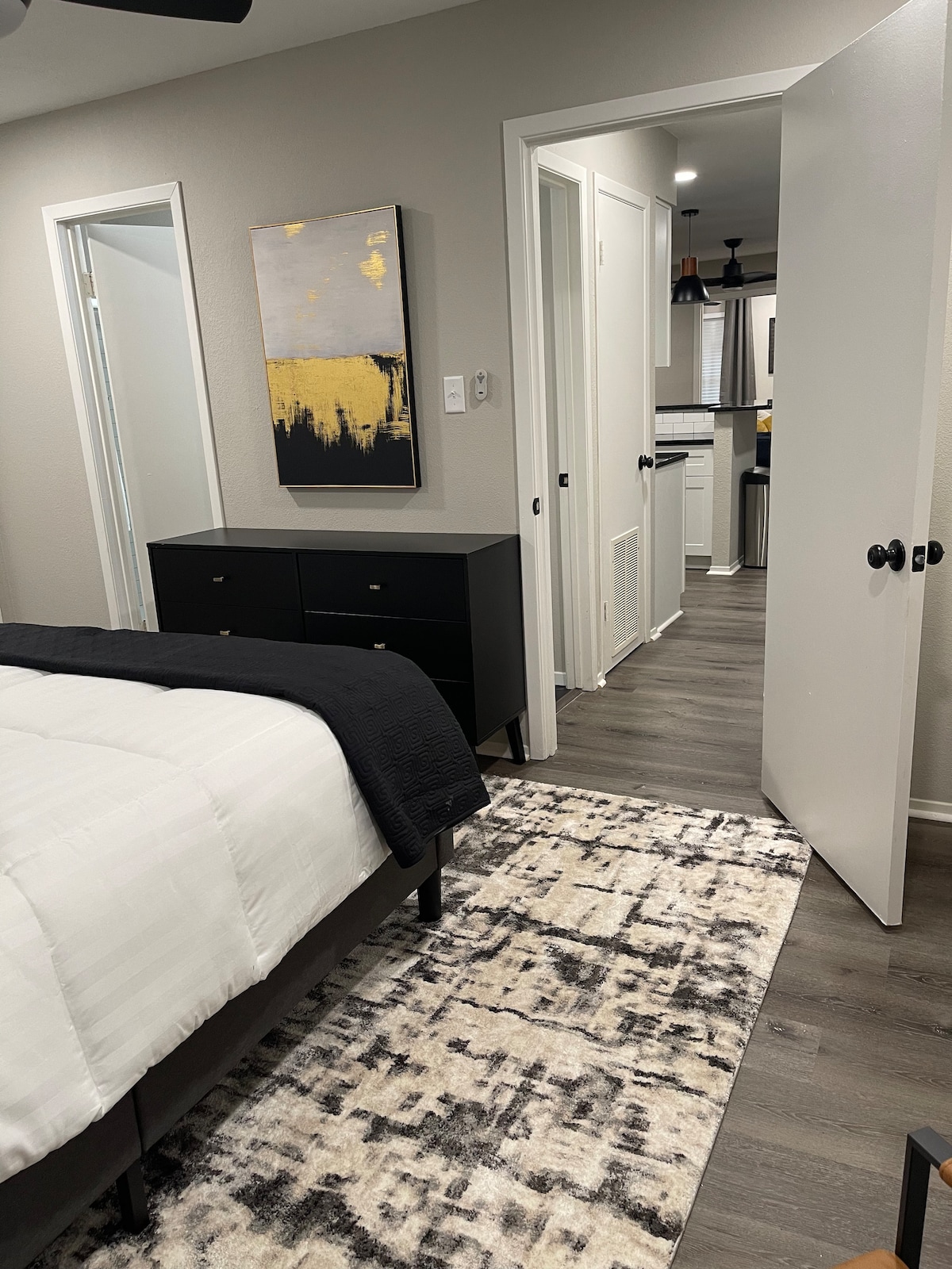The image depicts a modern bedroom featuring a spacious bed dressed in white linens. A black dresser stands against the wall, accompanied by abstract wall art above it. Light-colored doors lead to adjacent rooms, while a patterned area rug covers the floor.