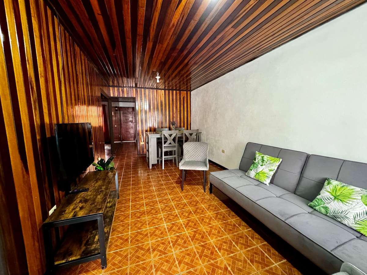 A living area features wooden paneling and a tiled floor. A gray sofa with decorative green pillows is positioned beside a television on a low wooden table. A dining area is visible in the background, with light-colored chairs contrasting the wood tones.
