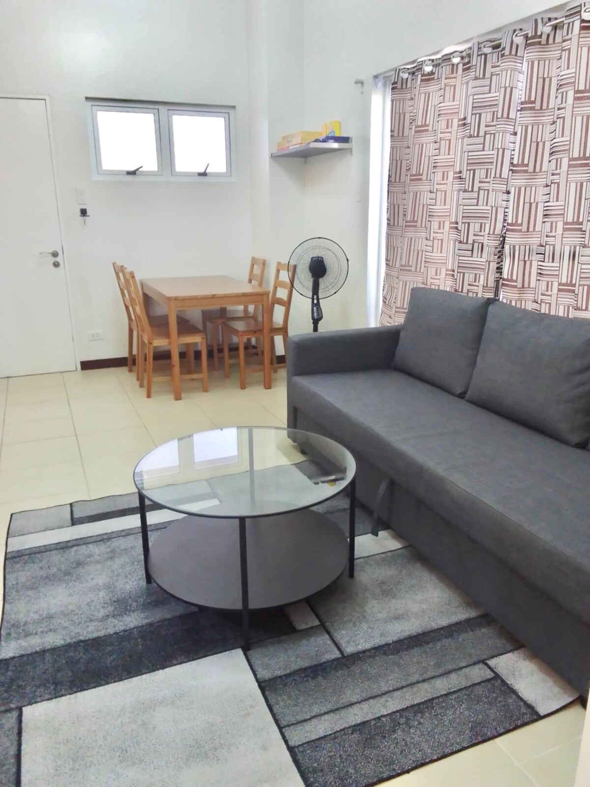 A living area showcases a modern IKEA sofabed in a muted gray shade. A circular glass coffee table sits on a geometric patterned rug. A four-seater dining area is positioned nearby, complemented by a wall-mounted shelf. Natural light filters through the window, partially covered by decorative curtains.