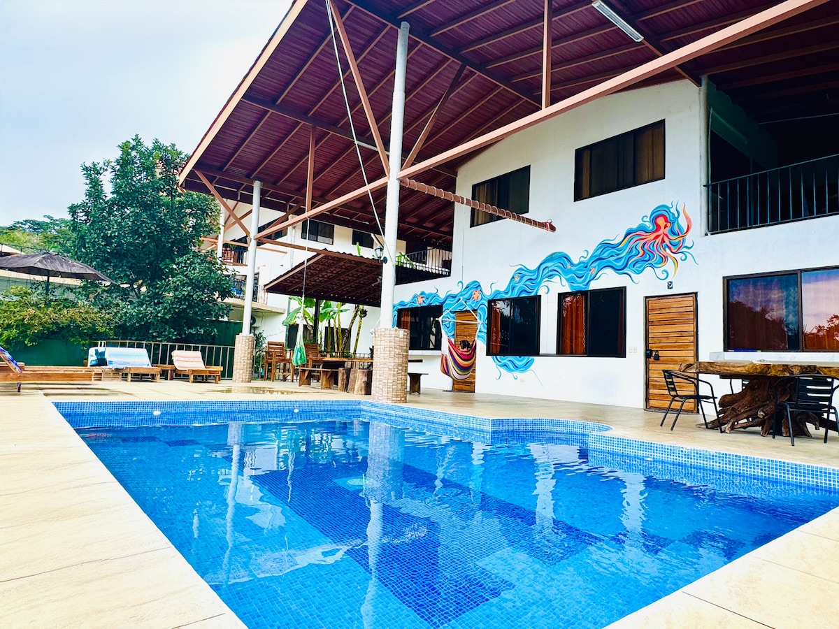 A tranquil pool featuring clear blue water reflects the surrounding greenery and architectural details of the multi-level building. Colorful mural artwork is visible across the exterior wall, while outdoor seating and dining areas are arranged under the canopy of the roof.