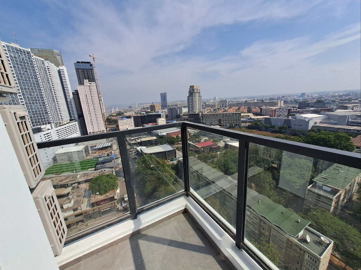A spacious balcony offers panoramic views of the urban landscape, showcasing a mix of residential and commercial buildings. The glass railing enhances the visibility of the surroundings, which includes distant views of the city skyline and a clear blue sky.