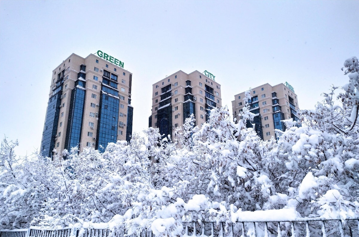 The exterior of the Green City Park residential complex is presented in a winter scene, with heavy snow covering the trees and pathways. The buildings are partially obscured by snow-laden branches, showcasing a modern architectural style with multiple balconies and large windows against a backdrop of a serene, white landscape.