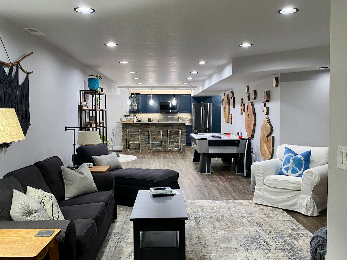 The living area features a cozy arrangement of a dark sofa, a chaise lounge, and an armchair, complemented by a sleek coffee table. The open kitchen is visible in the background, showcasing modern appliances and a counter with bar seating. Natural light is provided by recessed lighting and a window.