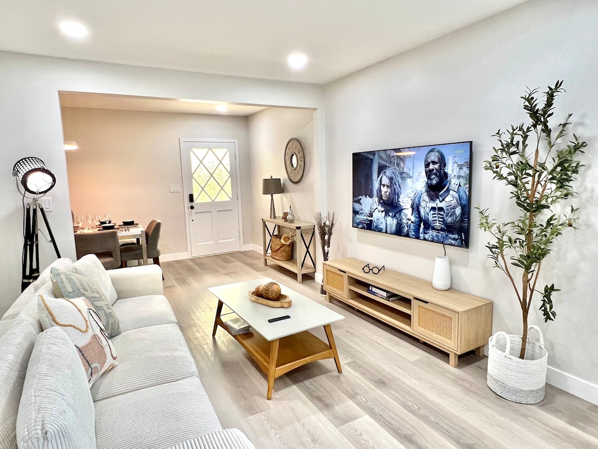 A cozy living area features a light-colored sectional sofa and a wooden coffee table with a decorative bowl. A smart TV is mounted on the wall opposite, while a dining area is visible through the doorway. Natural light streams in from the front door, enhancing the space.