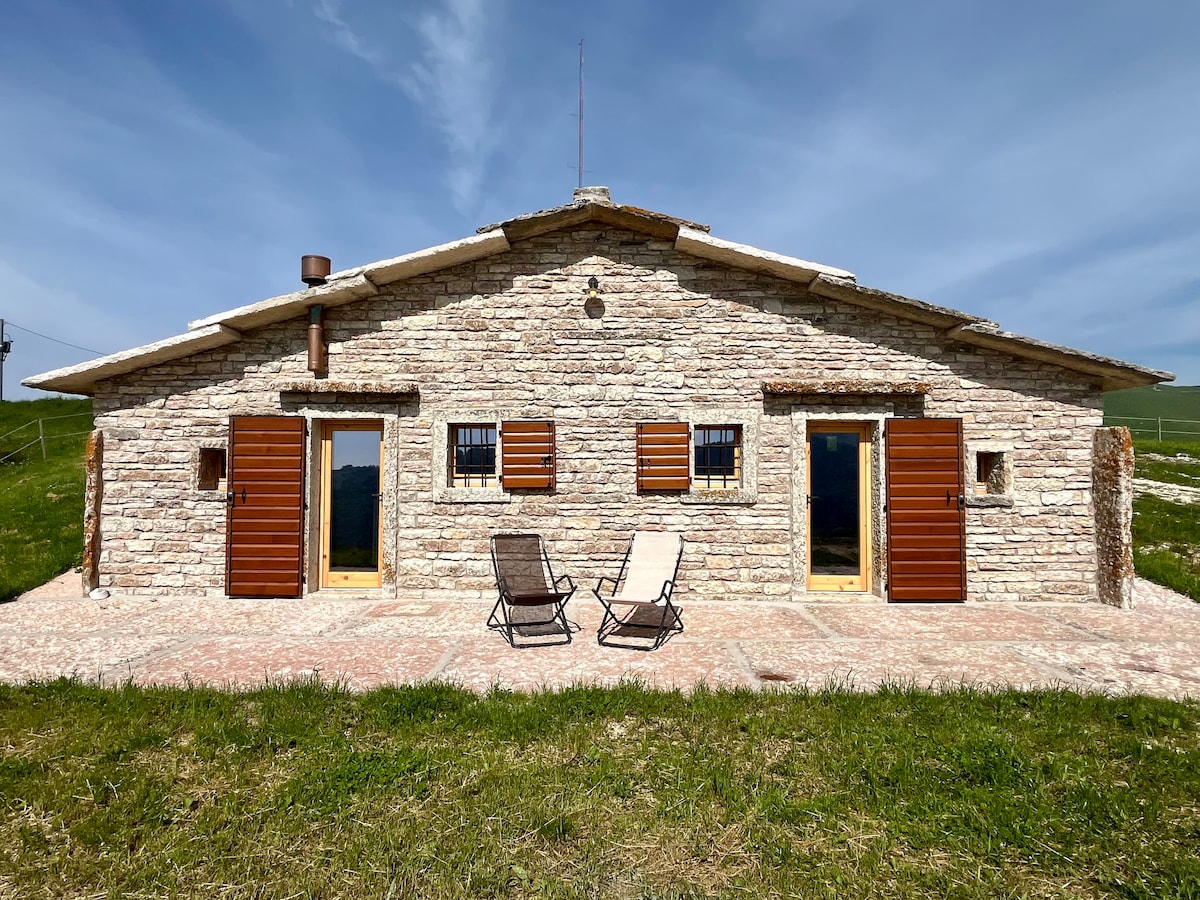 The chalet's exterior presents a charming stone façade, complemented by wooden shutters and doors. Two lounge chairs are placed on a spacious patio, surrounded by greenery, with mountains visible in the background under a clear blue sky.