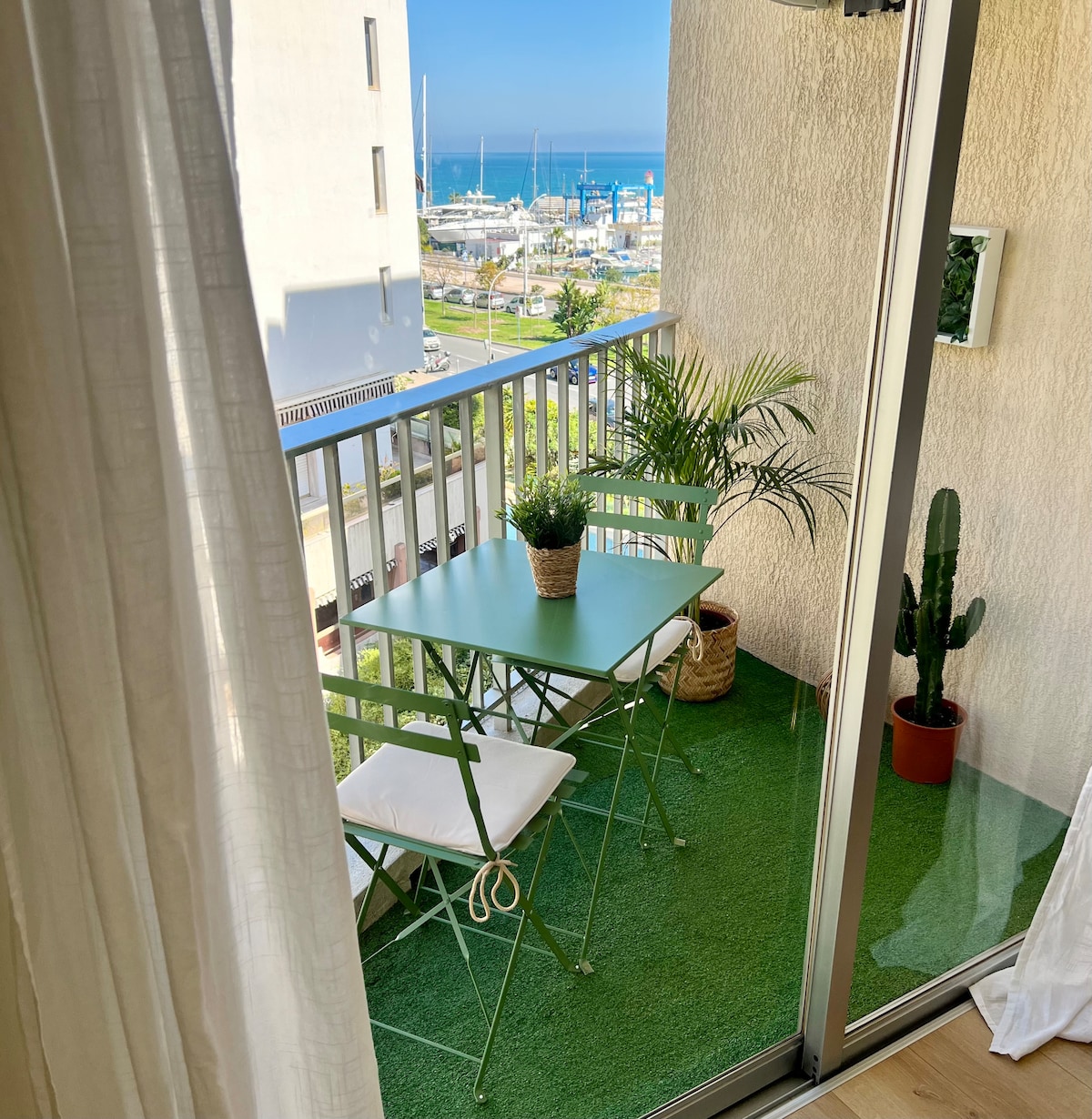 An inviting balcony features a green table with two folding chairs and potted plants. A view of the sea and marina is visible in the background, framed by a glass railing. Sunlight brightens the space, enhancing the vibrant colors of the plants.
