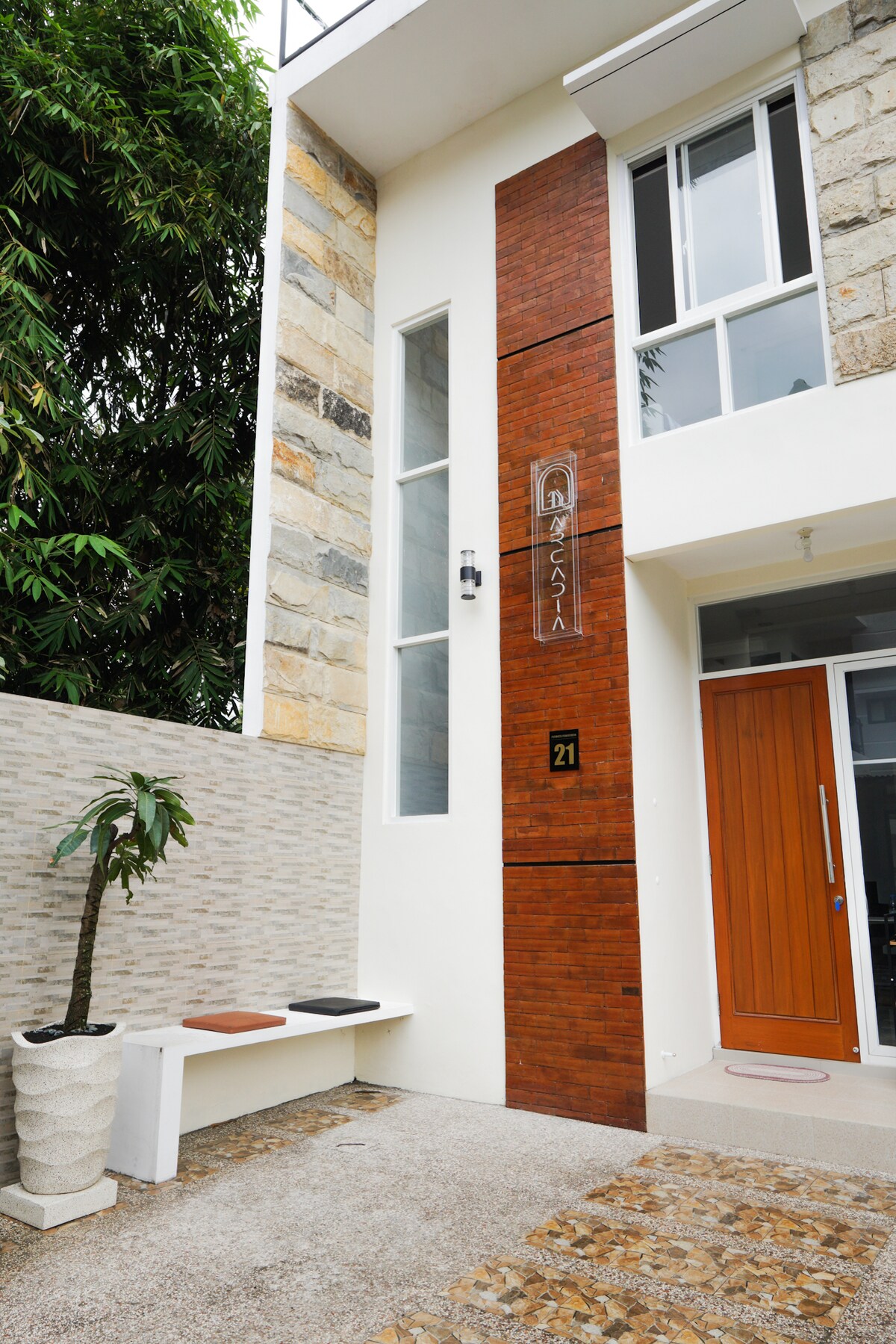 The entrance features a modern façade with a combination of textured stone and brick elements. A large window allows natural light to brighten the space. To the side, a minimalist seating area is included, adorned with a decorative plant in a white pot.