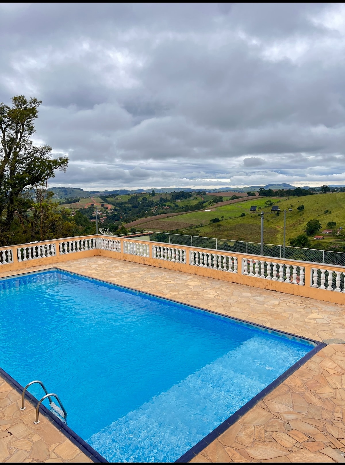 A clear blue swimming pool is framed by a stone patio and surrounded by a railing, offering views of rolling green hills under an overcast sky. The serene landscape is highlighted by patches of trees and distant hills.