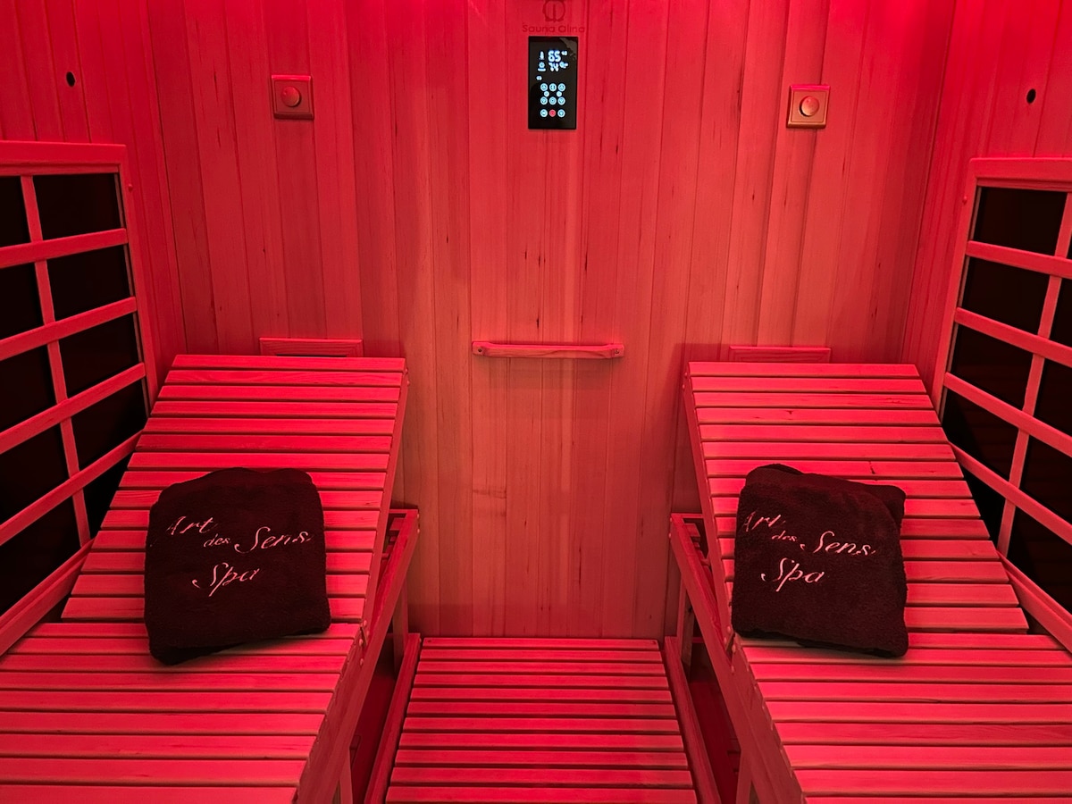 An infrared sauna room is presented with two spacious reclining benches made of wood. Each bench is adorned with a soft towel, and a digital control panel is visible on the wall, displaying the temperature settings. The room is illuminated with a red glow, creating a soothing atmosphere.