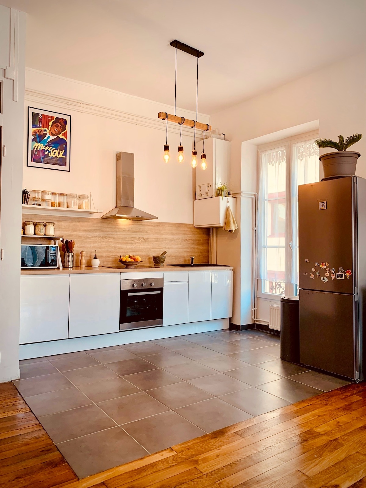 The kitchen features modern cabinetry with a white finish, a sleek stove, and an overhead light fixture with hanging bulbs. A large refrigerator stands against the wall, and natural light filters in through two adjacent windows. Countertop space includes decorative items and a fruit bowl.