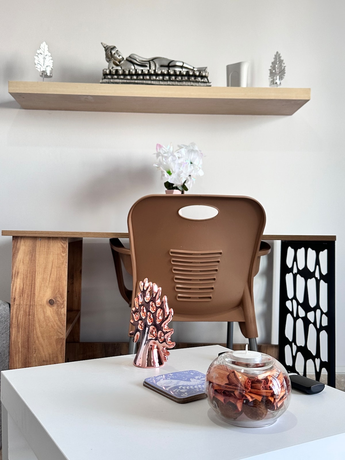A wooden work desk is complemented by a brown chair, positioned against a plain wall. A small shelf above holds decorative items, while a glass jar with colorful chocolates is placed on the white table in the foreground. An orchid plant adds a touch of nature.