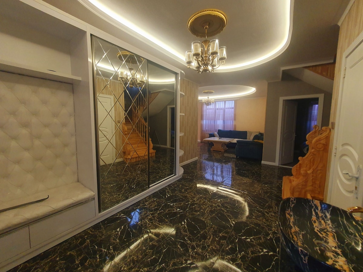 A spacious entryway is highlighted by a decorative chandelier and features polished dark marble flooring. A large mirror with a geometric pattern reflects the surroundings, while a cozy seating nook is lined with a tufted fabric. Soft, ambient lighting enhances the warm atmosphere.