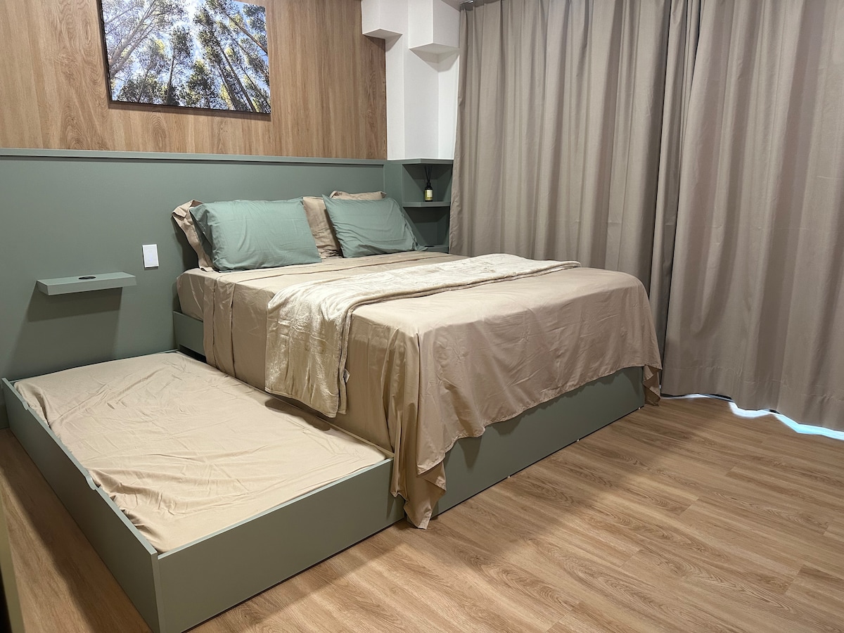 A sleeping area is designed with calming green walls and natural wood accents. A spacious bed is dressed in neutral-colored linens, complemented by soft pillows. Adjacent, an additional bed is neatly tucked beneath the main bed, providing extra sleeping space. The light wood flooring enhances the overall warmth.