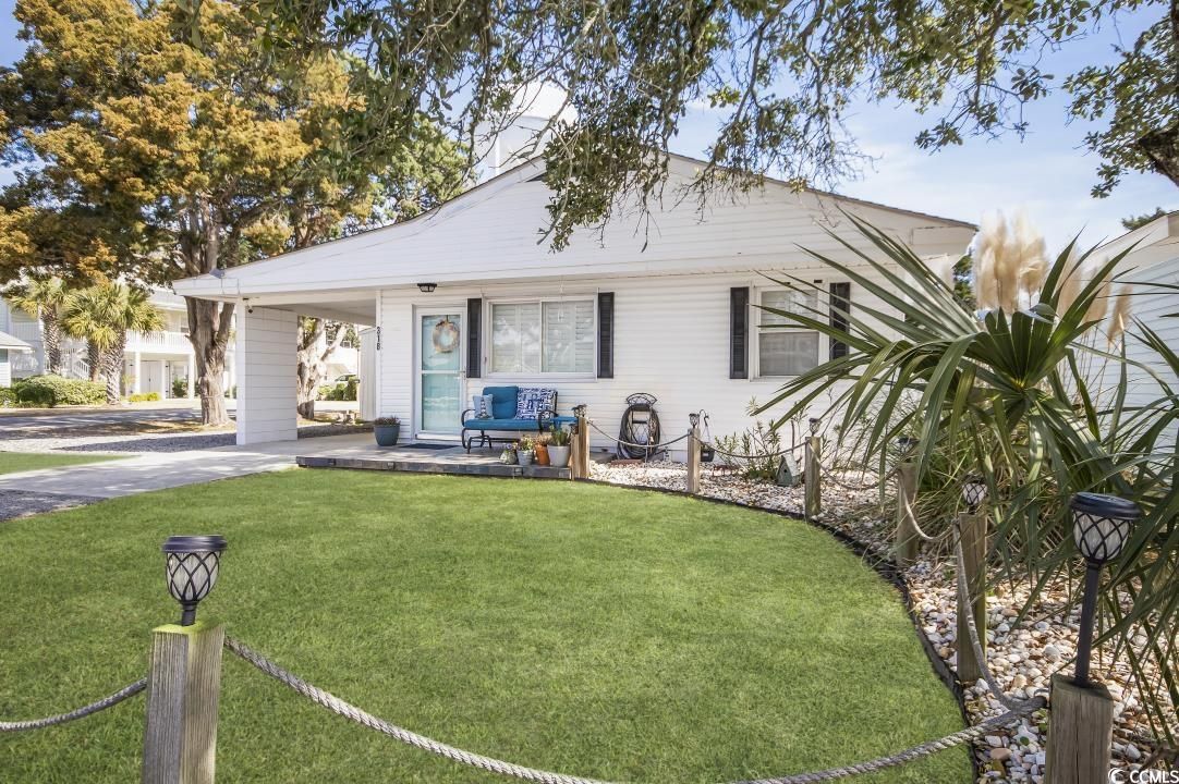 The exterior of a charming cottage is displayed, featuring a well-maintained front yard with green grass and decorative stones. A covered porch hosts blue seating, framed by lush greenery and trees. Soft lighting fixtures line the pathway, enhancing the inviting entrance.