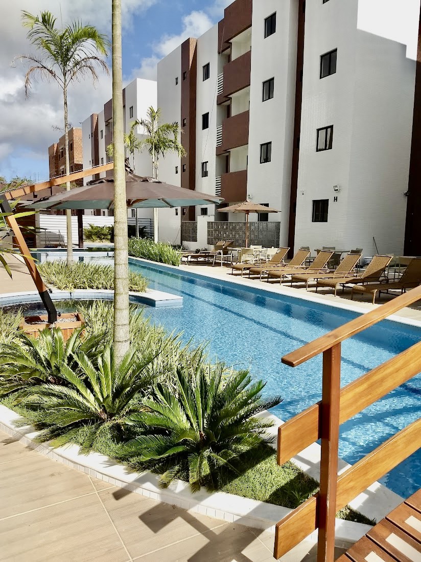 A swimming pool is surrounded by lounging chairs and shaded umbrellas, framed by well-maintained greenery. The building's modern architecture is visible in the background, with multiple stories and large windows reflecting natural light.
