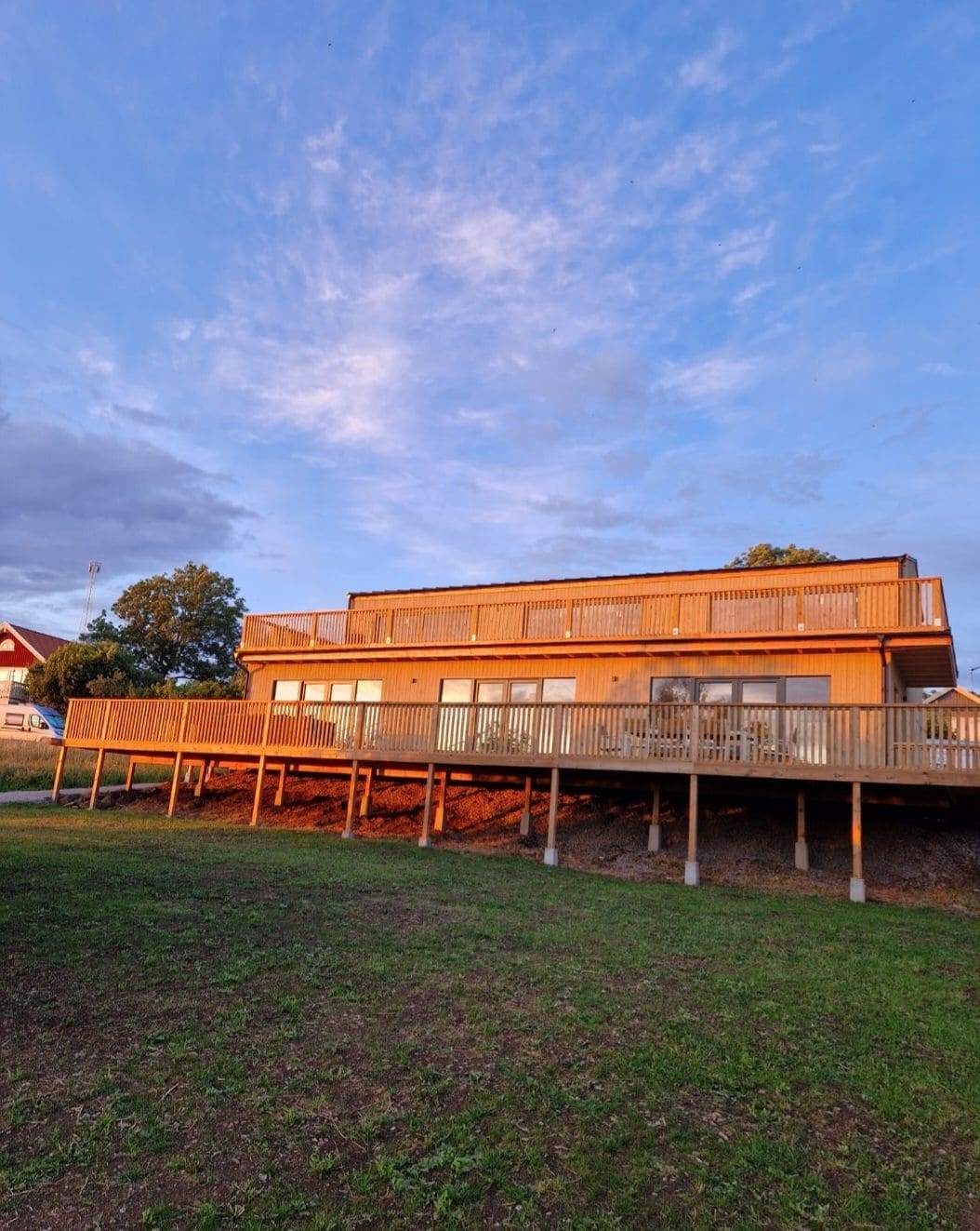 A two-story wooden villa is shown with a large deck that wraps around the front. The exterior features a light wood finish and large windows, allowing for natural light. A grassy area leads up to the structure, framed by trees under a partly cloudy sky.