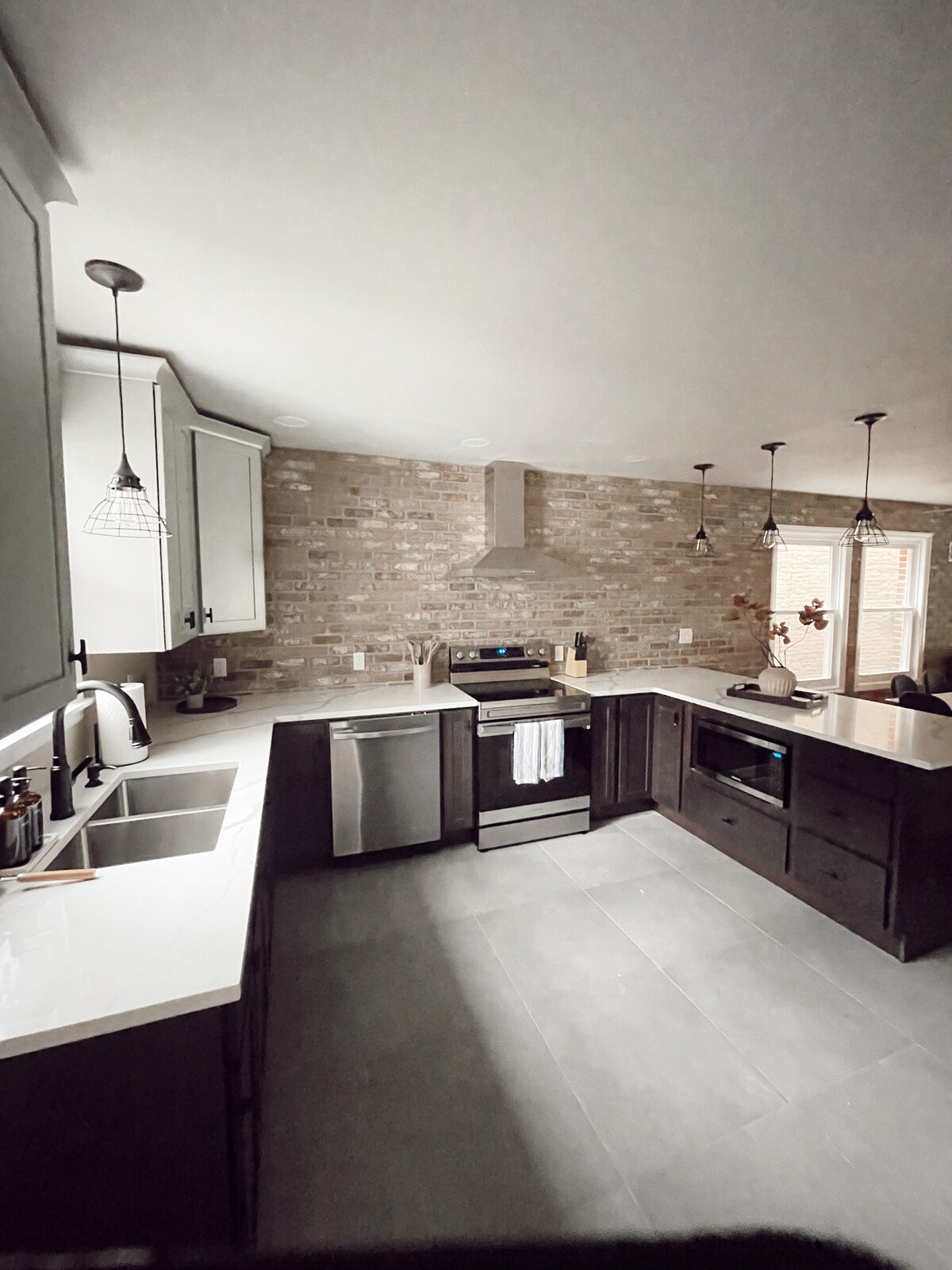 A modern kitchen is highlighted by dark cabinetry and white countertops. Stainless steel appliances include a refrigerator and an oven. A brick wall adds texture, while pendant lights provide illumination. The kitchen features a double sink and a window that allows for natural light.