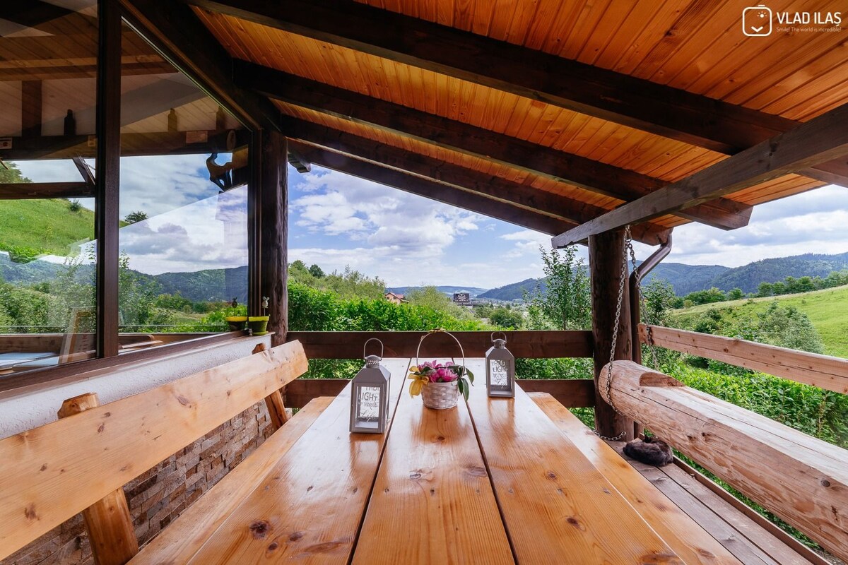 A wooden dining table with comfortable seating is set under a high, slanted ceiling, surrounded by large windows that frame a scenic view of rolling hills and greenery. Two decorative lanterns and a flower arrangement are positioned on the table.