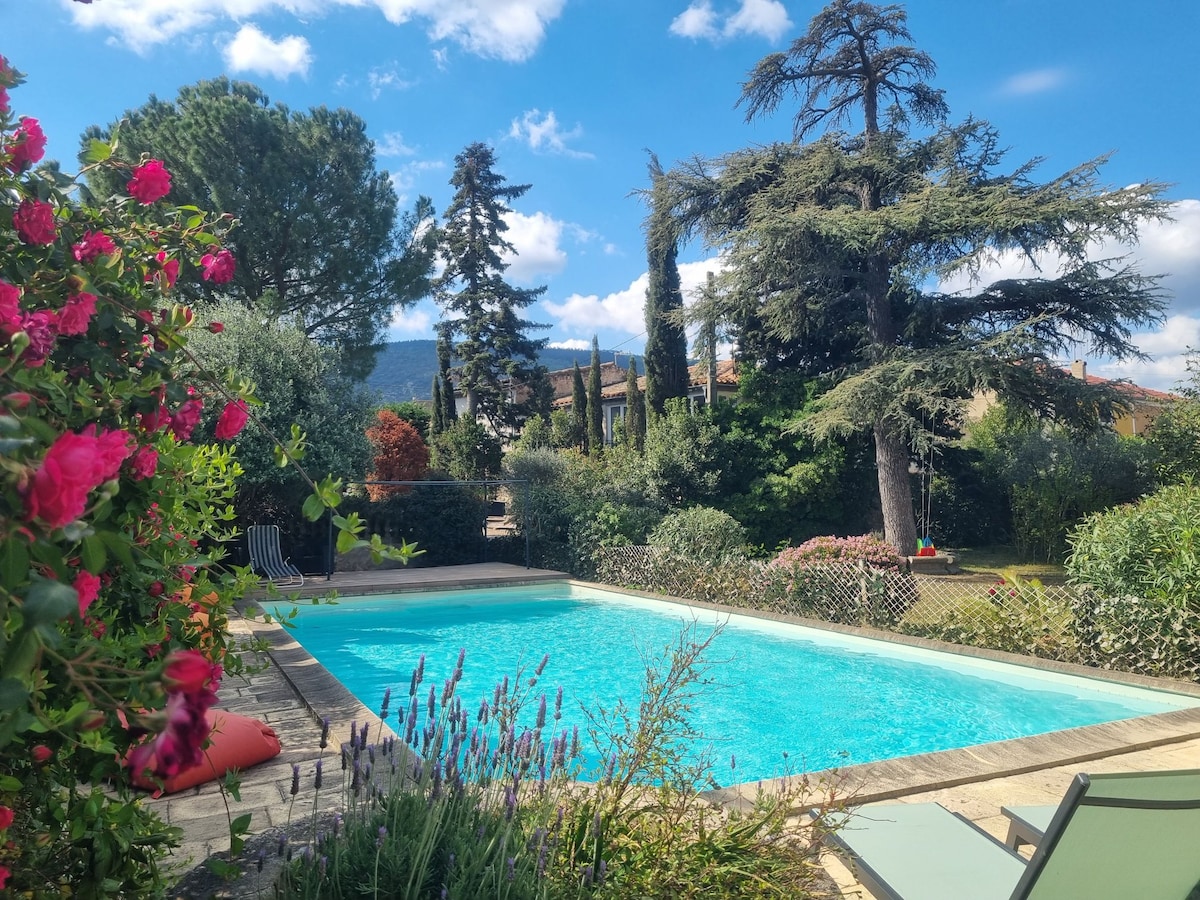 A serene outdoor pool area is surrounded by vibrant flowers and lush greenery. Tall trees provide shade, while the clear blue water reflects the sky. Lounge chairs are positioned nearby, inviting relaxation in a tranquil setting.