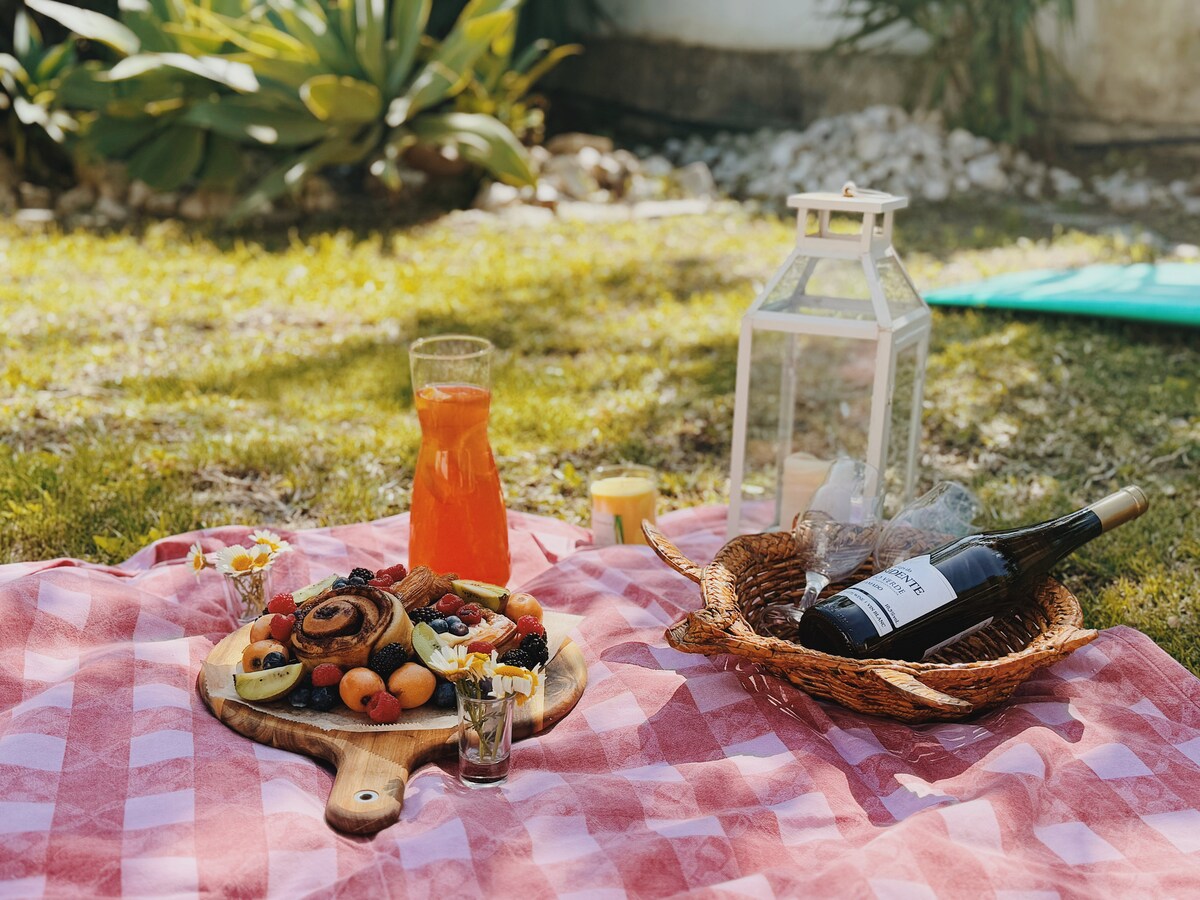 A picnic setup is seen on a checkered blanket spread across the grass. A wooden platter displays a variety of fruits and pastries. A glass pitcher holds a colorful beverage, alongside a bottle of wine and a lantern, adding a serene atmosphere to the outdoor space.