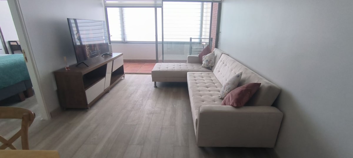 A light-filled living area features a modern beige sectional sofa adorned with two decorative pillows. A flat-screen TV is positioned on a wooden media console. Large windows provide a view of the outside, bringing in natural light and a feeling of spaciousness.