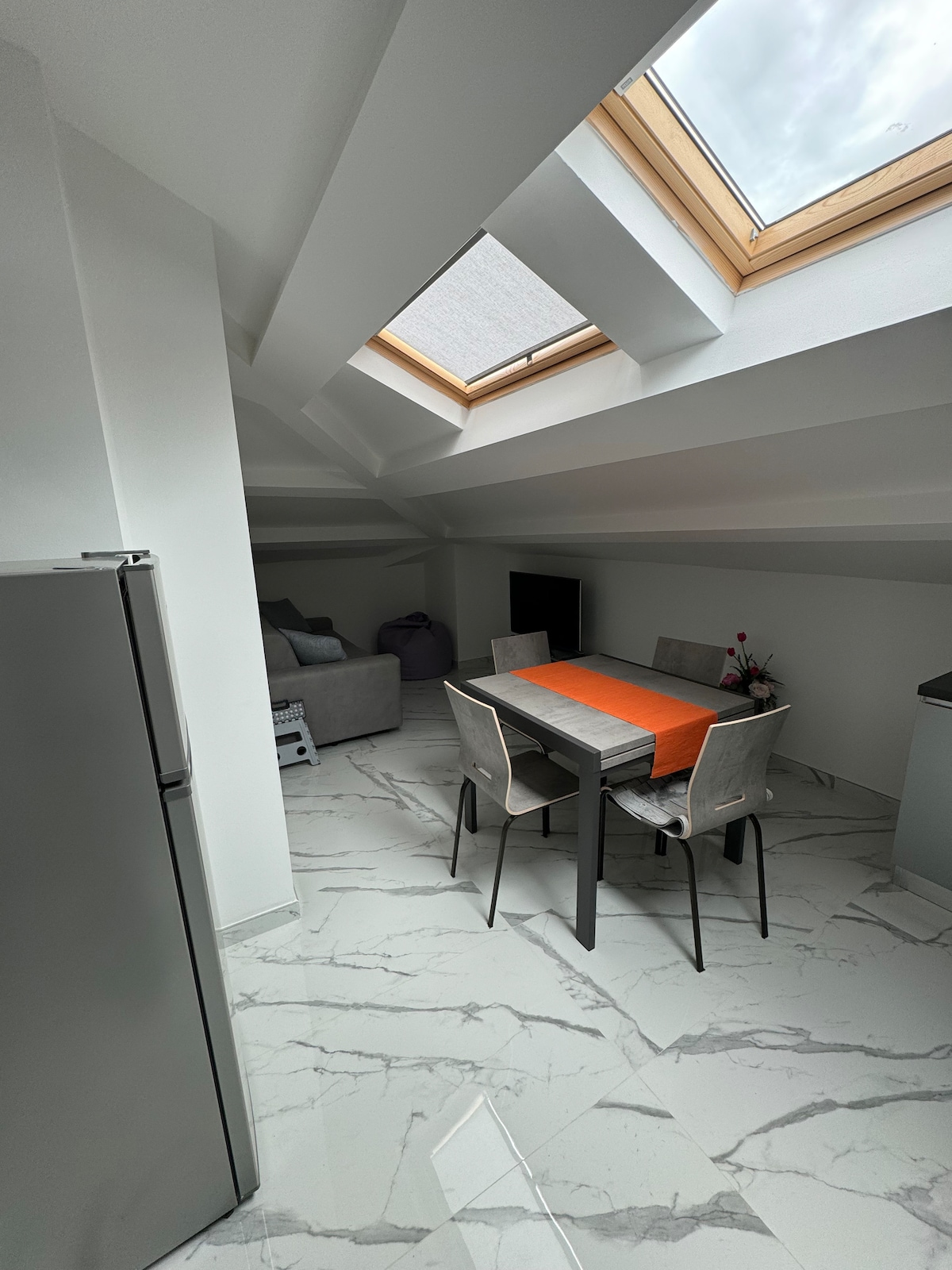 An airy dining area is illuminated by natural light from a skylight above. A rectangular table is set with an orange table runner, surrounded by four light gray chairs. A cozy seating arrangement with a soft sofa is visible in the background, contributing to a comfortable atmosphere.