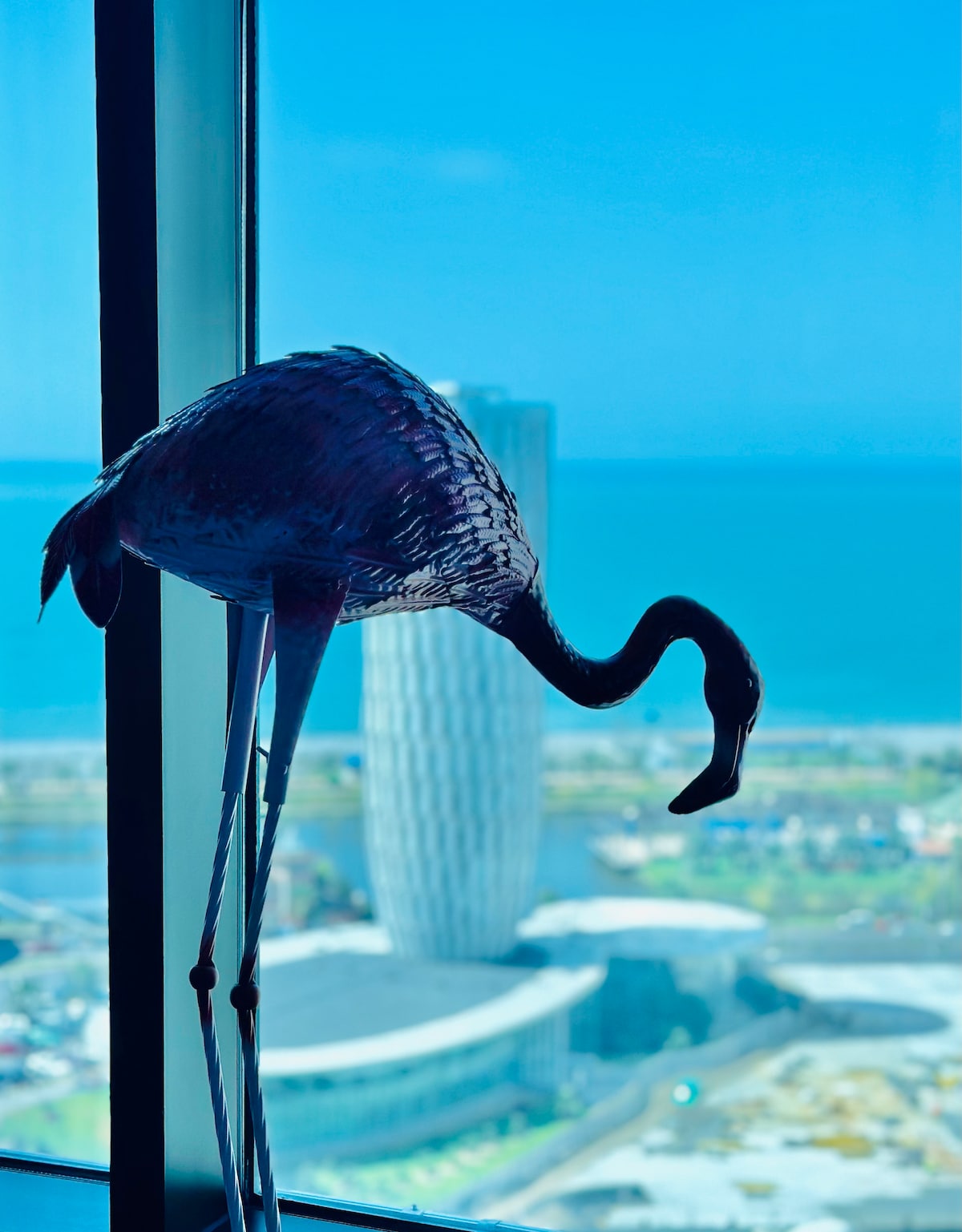 A decorative flamingo stands gracefully by the window, complementing the panoramic view of the sea and the skyline of nearby buildings. The striking blue of the water enhances the tranquility of the scene, while the modern architecture of the skyscrapers can be seen in the background.