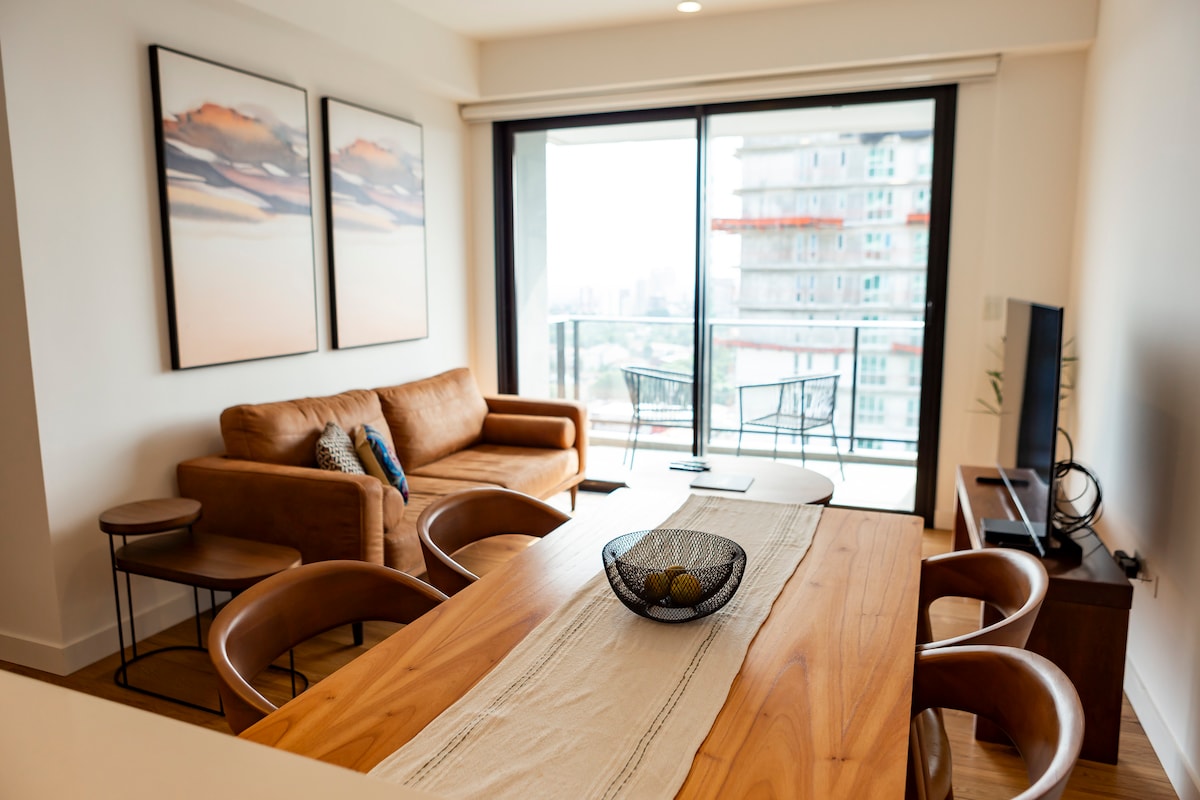 A modern living space features a wooden dining table surrounded by curved chairs. A stylish sofa sits nearby, complemented by abstract artwork on the walls. Large glass doors lead to a balcony, allowing natural light to fill the room.